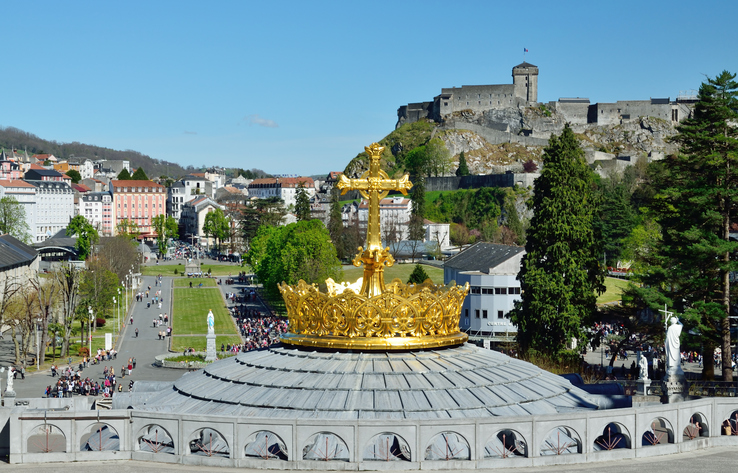Lourdes is a major place of Roman Catholic pilgrimage. There is a dome of the Rosary basilica surmounted with the gilded crown and the cross in the foreground.