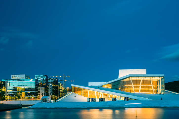 Oslo, Norway - July 31, 2014: The Scenic Night Evening View Of Illuminated Norwegian National Opera And Ballet House Among Contemporary High-Rise Buildings. Blue Sky Background, Copyspace.