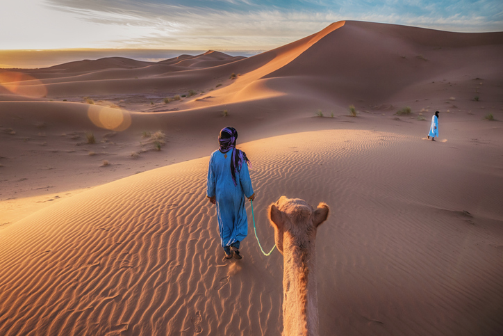 Two Muslim men dressed in blue robes wearing headscarves walking through the sand dunes of the Sahara Desert, leading a camel at sunrise, with golden light and lens flare.