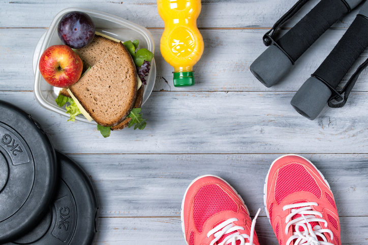 Fitness concept, pink sneakers, weight plates, dumbbells, sandwich, fruits and orange juice, wooden background, top view
