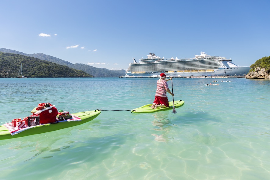 Oasis of the Seas, holiday, rear view of Santa kneeling on paddleboard, paddle board, rowing, towing another board behind with his bag of gifts, presents on a beach towel, paddling toward guests swimming near Oasis of the Seas at Labadee beach, Haiti, fun