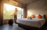 Portrait of tourist woman standing nearly window, looking to beautiful view with her luggage in hotel bedroom after check-in.