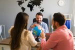 Agências e recomendações para quem pretende fazer intercâmbio vista-traseira-de-uma-jovem-hispanica-e-um-homem-olhando-para-paises-em-um-globo-enquanto-planeja-sua-viagem-ao-exterior-com-um-agente-de-viagens